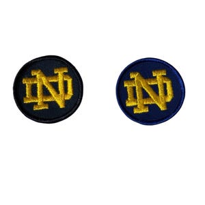 May include: Two embroidered patches with the Notre Dame Fighting Irish logo. The logo is gold and the patches are circular. One patch is black and the other is blue.