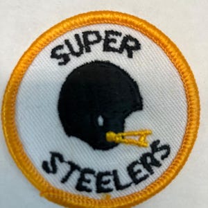 Peut inclure: Un écusson circulaire blanc et orange avec les mots "SUPER STEELERS" brodés en noir. Une silhouette noire d'un casque de football américain avec une visière dorée est au centre.