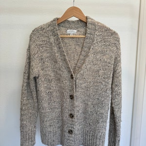 Puede incluir: Un cárdigan beige con cuello en V y cierre de botones. El cárdigan está hecho de un tejido de punto suave y texturizado.