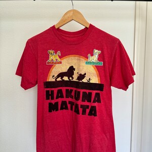 May include: Red t-shirt with a graphic of Simba, Timon, and Pumbaa from The Lion King. The graphic includes a sunset and the text "Hakuna Matata".