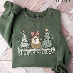 May include: Military green sweatshirt with a Christmas-themed graphic. The design features a nativity scene with two Christmas trees and the text "Let Heaven Nature Sing". The sweatshirt is made of a soft material.