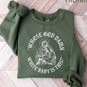 May include: Military green sweatshirt with white text and a religious illustration. The text reads "WHOS GOD DAMN WHITE BABY IS THIS?" in a circular design around the illustration of a religious figure holding a baby.