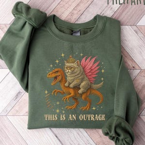 May include: Military green sweatshirt featuring a whimsical graphic of a cat wearing glasses and a crown, riding a dinosaur with pink wings. The text "THIS IS AN OUTRAGE" is printed below the image. The sweatshirt has long sleeves.