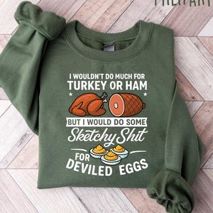 May include: Military green sweatshirt with the text "I wouldn't do much for turkey or ham but I would do some sketchy shit for deviled eggs." The graphic includes illustrations of a roasted turkey, ham, and deviled eggs.
