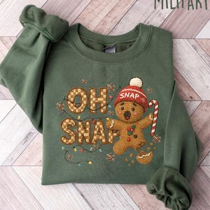 May include: Military green sweatshirt with the text "OH SNAP" and a gingerbread man wearing a red hat and holding a candy cane. The design includes string lights and a small bowl of crumbs. The sweatshirt is folded on a light wood surface.