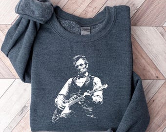 May include: Heather dark gray sweatshirt featuring a white graphic of Abraham Lincoln playing a guitar. The image shows a detailed illustration of Lincoln in a vest and shirt, holding the guitar. The sweatshirt is folded, with the text "HEATHER DARK GRAY" at the top.