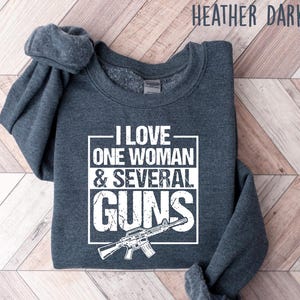 May include: Heather dark gray sweatshirt with the text "I LOVE ONE WOMAN & SEVERAL GUNS" in white. A graphic of a rifle is below the text. The sweatshirt is folded on a wooden surface.