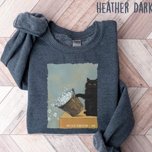 May include: Heather dark gray sweatshirt featuring a whimsical illustration of a black cat with a bucket of ice. The cat is standing next to the bucket, with ice spilling out. The text "NO ICE VERSION 1888" is printed on the bottom.
