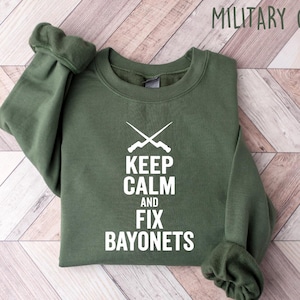May include: Military green sweatshirt with the text "KEEP CALM AND FIX BAYONETS" in white. The design includes crossed bayonets above the text. The sweatshirt is folded on a light wood-grain surface.