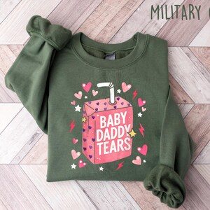 May include: Military green sweatshirt with a pink juice box graphic that reads "BABY DADDY TEARS". The graphic includes pink hearts, stars, and lightning bolts. The sweatshirt is folded on a light wood-grain surface.