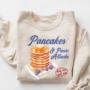 May include: A beige sweatshirt with a graphic of a stack of pancakes with a pat of butter on top and blueberries. The text "Pancakes & Panic Attacks" is printed in blue above the pancakes. The text "buy one get one" is printed in red below the pancakes.