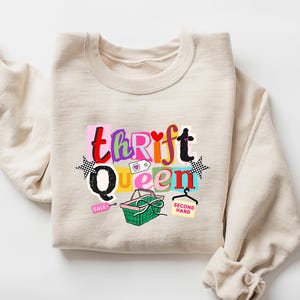 May include: A sand-colored sweatshirt with the text "thrift Queen" in colorful, playful lettering. The design includes a shopping basket, stars, and the words "sale" and "second hand". The sweatshirt is folded with the sleeves rolled up.