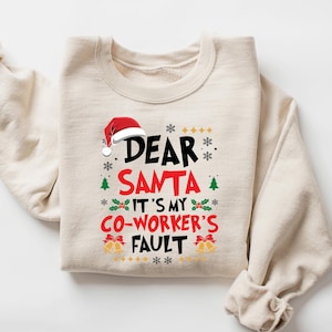 May include: A sand-colored sweatshirt with the text "DEAR SANTA IT'S MY CO-WORKER'S FAULT" in a festive design. The design includes a Santa hat, snowflakes, Christmas trees, and bells, perfect for the holiday season.