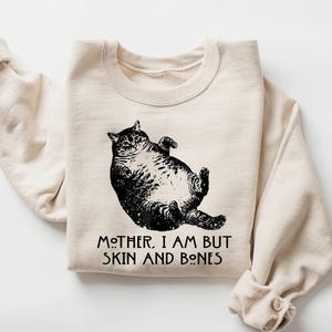 May include: A sand-colored sweatshirt with a black graphic of a cat lying on its back. The text "MOTHER, I AM BUT SKIN AND BONES" is printed below the cat. The sleeves are folded.