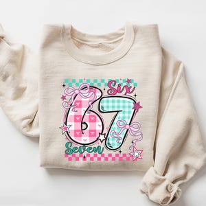 May include: A beige sweatshirt with a colorful graphic design. The design features the numbers "67" in pink and teal, with the words "Six" and "Seven" above and below. The numbers are decorated with bows and stars, set against a checkered background.