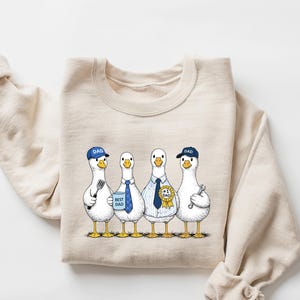 May include: A sand-colored sweatshirt featuring a whimsical illustration of four geese dressed as dads. Each goose wears a hat or tie and holds a tool or sign. The text "DAD" is on the hats and "BEST DAD" on a sign.