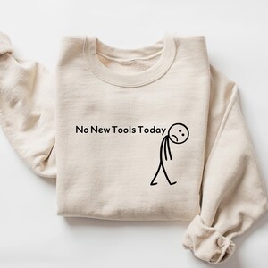 May include: A sand-colored sweatshirt with the text "No New Tools Today" and a stick figure with a sad face. The sweatshirt is made of a soft material and has a crew neck.