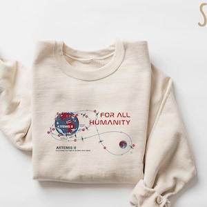 May include: A sand-colored sweatshirt with the text "FOR ALL HUMANITY" in red. The graphic depicts a stylized map of the Artemis II mission, with a blue and red Earth and a dotted flight path.