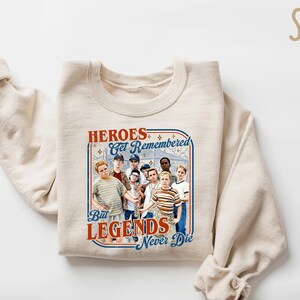 May include: A sand-colored sweatshirt with the text "HEROES Get Remembered But LEGENDS Never Die" in a retro design. The graphic features a group of boys. The sweatshirt is folded with the sleeves rolled up.