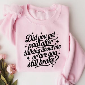 May include: A light pink sweatshirt with the text "Did you get paid after talking about me or are you still broke?" in black script. The sweatshirt is folded, and a bouquet of pink flowers is in the lower left corner.