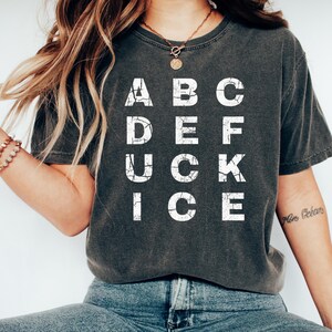May include: Dark gray t-shirt with the word "ABC DEF UCK ICE" in white, distressed lettering. The shirt is worn with blue jeans and white slip-on shoes. A gold necklace and bracelets are also visible.