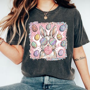 May include: Dark gray t-shirt featuring a graphic of a bunny wearing sunglasses and blowing a bubblegum bubble. The bunny is surrounded by Easter eggs on a pink background. The shirt is paired with blue jeans and white shoes.