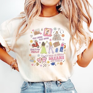 May include: A cream-colored short-sleeve t-shirt featuring a colorful graphic print with various illustrations including a red scooter, a suitcase, musical notes, flowers, and text that reads Hey Now Hey Now, Sing to me Paolo, and This is what dreams are made of.