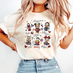 May include: Cream-colored t-shirt featuring a colorful graphic of Mickey Mouse and friends, including Pluto, Goofy, Donald Duck, and Minnie Mouse. The design includes the text "Mickey Mouse" and character signatures. The shirt is paired with light blue jeans.