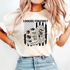 May include: Cream-colored t-shirt with a graphic featuring a Bigfoot holding a 10mm cartridge, set against a black and white American flag design. The text "LOOKING FOR THIS?" is printed above the graphic.