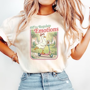 May include: Cream-colored t-shirt featuring a graphic of a goose riding a pink scooter, holding a coffee cup, and wearing a bow. The graphic is framed with the text "Off to Regulate Emotions". The shirt is paired with blue jeans.