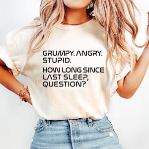 May include: Cream-colored t-shirt with the text "GRUMPY. ANGRY. STUPID. HOW LONG SINCE LAST SLEEP, QUESTION?" in black. The shirt is paired with light blue denim jeans.