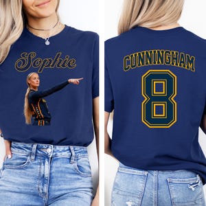 May include: A navy blue t-shirt featuring a graphic of a basketball player pointing, the name Sophie in script, and the back displaying the name CUNNINGHAM with the number 8 in a bold yellow and navy blue athletic font style on the back.