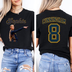 May include: A black short-sleeved t-shirt featuring a graphic on the front with the name Sophie in yellow script above a photo of a basketball player. The back of the shirt displays the name CUNNINGHAM in yellow block letters above the number 8.