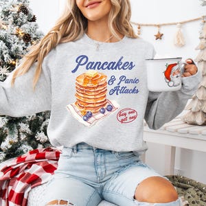 Op de afbeelding: Een grijs sweatshirt met een afbeelding van een stapel pannenkoeken en de tekst "Pancakes & Panic Attacks" op een blauw-wit geruite achtergrond. De tekst "Buy one get one" is ook in de afbeelding opgenomen.