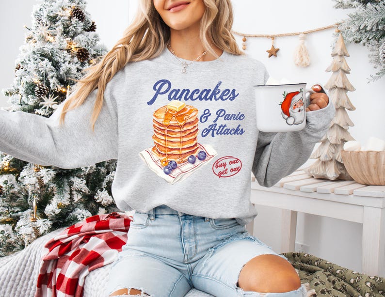 Op de afbeelding: Een grijs sweatshirt met een afbeelding van een stapel pannenkoeken en de tekst "Pancakes & Panic Attacks" op een blauw-wit geruite achtergrond. De tekst "Buy one get one" is ook in de afbeelding opgenomen.