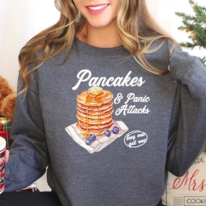 Op de afbeelding: Een grijs sweatshirt met een witte afbeelding van pannenkoeken en de tekst "Pancakes & Panic Attacks" en "buy one get one".