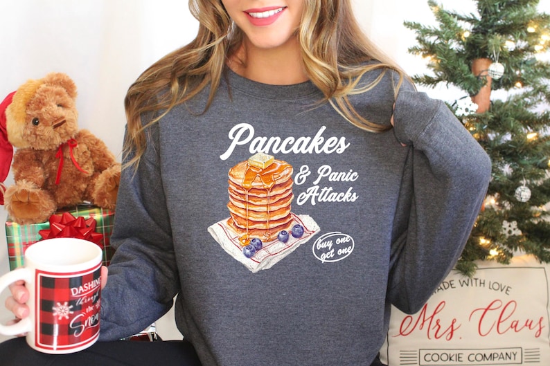 Op de afbeelding: Een grijs sweatshirt met een witte afbeelding van pannenkoeken en de tekst "Pancakes & Panic Attacks" en "buy one get one".