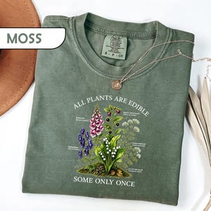 May include: A moss green short-sleeve t-shirt featuring a botanical illustration of various plants with the text "ALL PLANTS ARE EDIBLE SOME ONLY ONCE". The graphic includes labels for foxglove, monkshood, deadly nightshade, and lily of the valley.