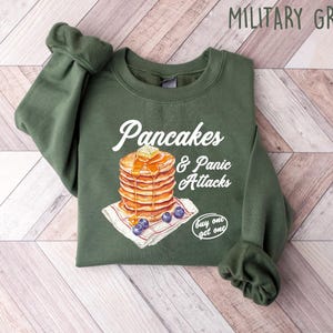 Op de afbeelding: Een groen sweatshirt met de tekst "Pancakes & Panic Attacks" en een afbeelding van een stapel pannenkoeken met blauwe bessen en een "koop er &eacute;&eacute;n, krijg er &eacute;&eacute;n gratis"-tag.