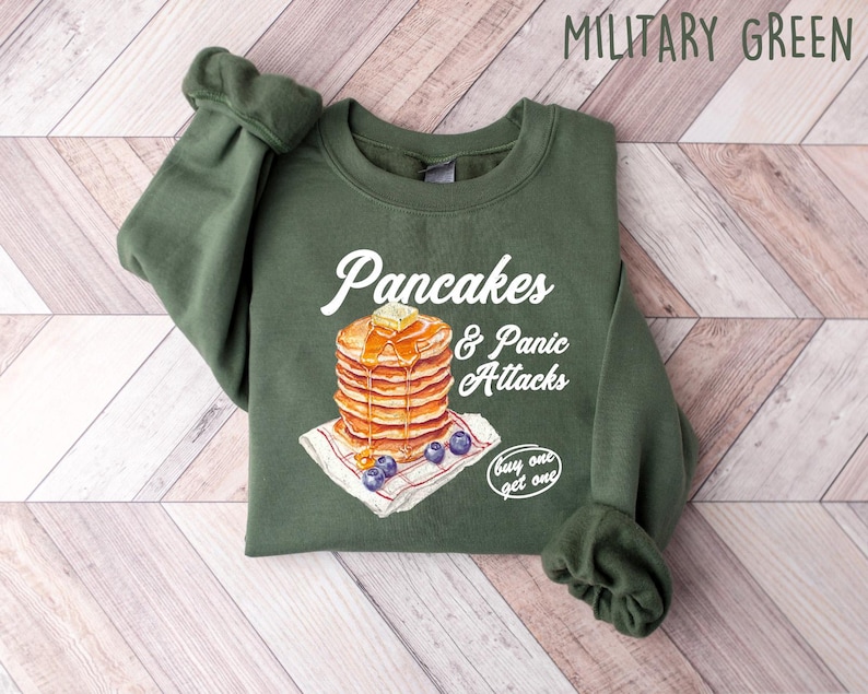 Op de afbeelding: Een groen sweatshirt met de tekst "Pancakes & Panic Attacks" en een afbeelding van een stapel pannenkoeken met blauwe bessen en een "koop er &eacute;&eacute;n, krijg er &eacute;&eacute;n gratis"-tag.