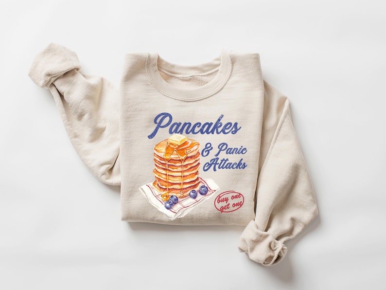 Op de afbeelding: Een beige sweatshirt met een afbeelding van een stapel pannenkoeken met een klontje boter erop en blauwe bessen. De tekst "Pancakes & Panic Attacks" is in blauw boven de pannenkoeken gedrukt. De tekst "buy one get one" is in rood onder de pannenkoeken gedrukt.