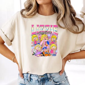 May include: A cream-colored short-sleeve t-shirt featuring a graphic print of the cartoon character Lizzie McGuire in various poses. The design includes the text "LIZZIE McGuire" in a rainbow-colored font, surrounded by flowers and lightning.