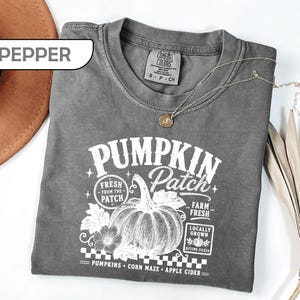 May include: A pepper-colored short-sleeve t-shirt featuring a white graphic print of a pumpkin with the text PUMPKIN Patch, Fresh From The Patch, Farm Fresh, Locally Grown, Autumn Picked, Pumpkins, Corn Maze, and Apple Cider on the front.