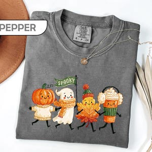 May include: A pepper-colored t-shirt featuring a graphic of a pumpkin, a ghost holding a green flag with the text SPOOKY, an autumn leaf, and a coffee cup, all with smiling faces. The shirt has a crew neckline and a Comfort Colors brand label.