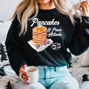Op de afbeelding: Zwart sweatshirt met een witte afbeelding van een stapel pannenkoeken met blauwe bessen en de tekst "Pancakes & Panic Attacks buy one get one".