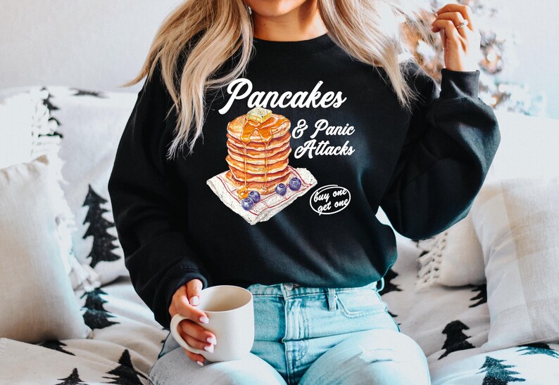 Op de afbeelding: Zwart sweatshirt met een witte afbeelding van een stapel pannenkoeken met blauwe bessen en de tekst "Pancakes & Panic Attacks buy one get one".