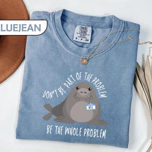 May include: A blue jean colored t-shirt featuring a graphic of a grey seal wearing a name tag that says "NEIL". The text above the seal reads "DON'T BE PART OF THE PROBLEM" and the text below the seal reads "BE THE WHOLE PROBLEM". The shirt has a crew neckline and a Comfort Colors brand label.