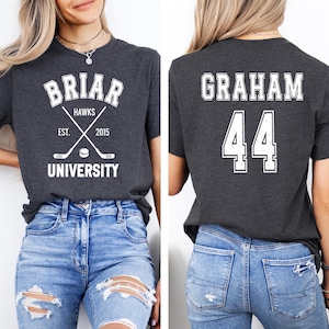 May include: A dark gray short-sleeve t-shirt featuring white graphics. The front displays the text BRIAR HAWKS EST. 2015 UNIVERSITY with crossed hockey sticks and a puck. The back features the name GRAHAM and the number 44 in a collegiate font.