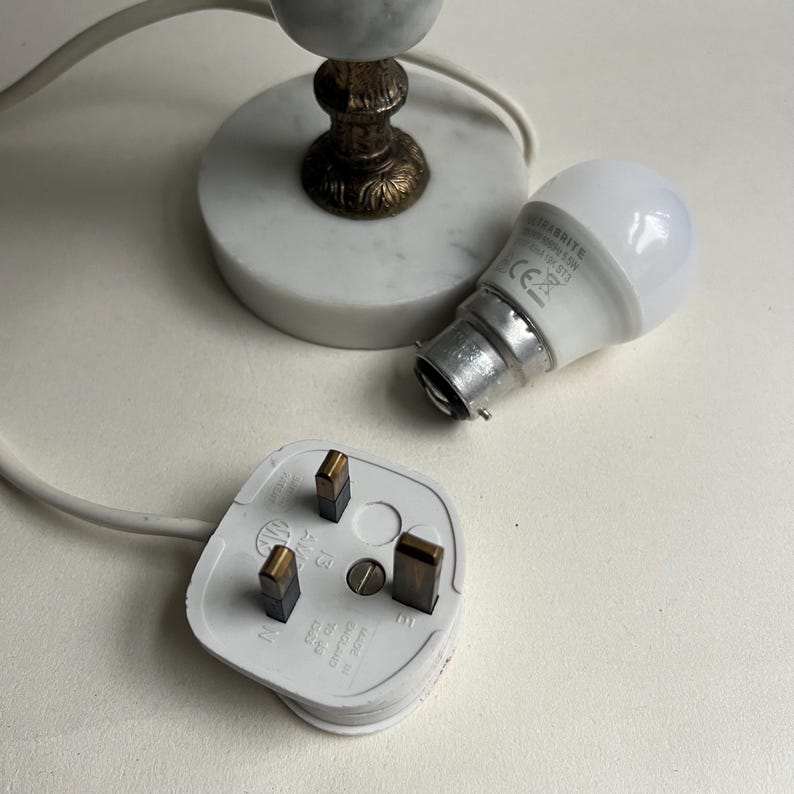 May include: A white electrical plug with three prongs, a white cord, and a light bulb with a silver base. A small lamp base with a marble top and ornate brass stem is also visible.