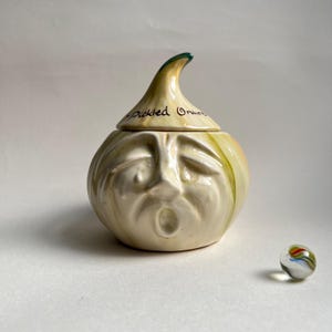 May include: A ceramic jar shaped like an onion with a face, labeled "Pickled Onions." The jar is off-white with green and yellow accents. A small, colorful marble sits nearby. The jar has a lid with a pointed top.